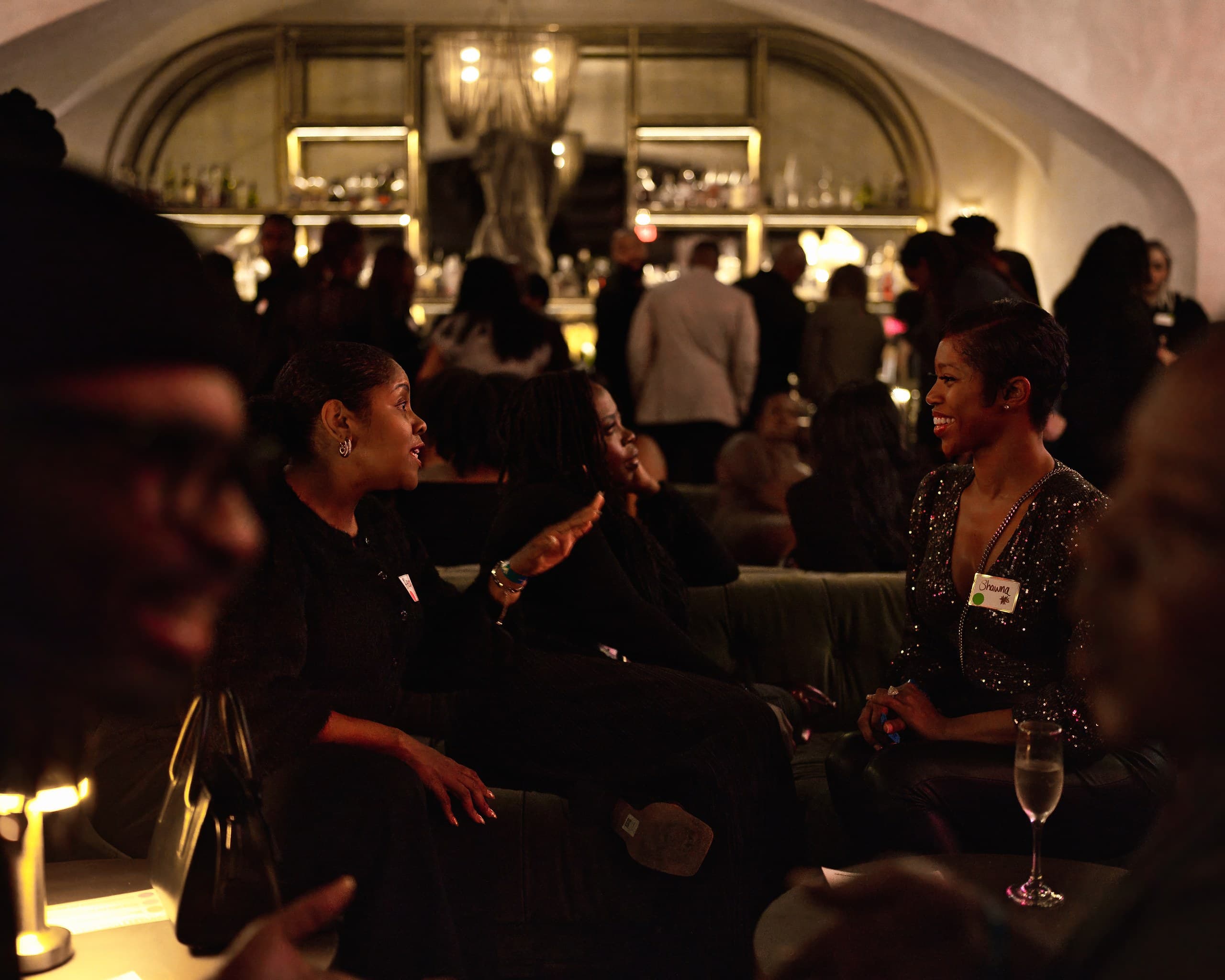 Guests talking on velvet sofas in a candlelit lounge with a glowing bar behind them
