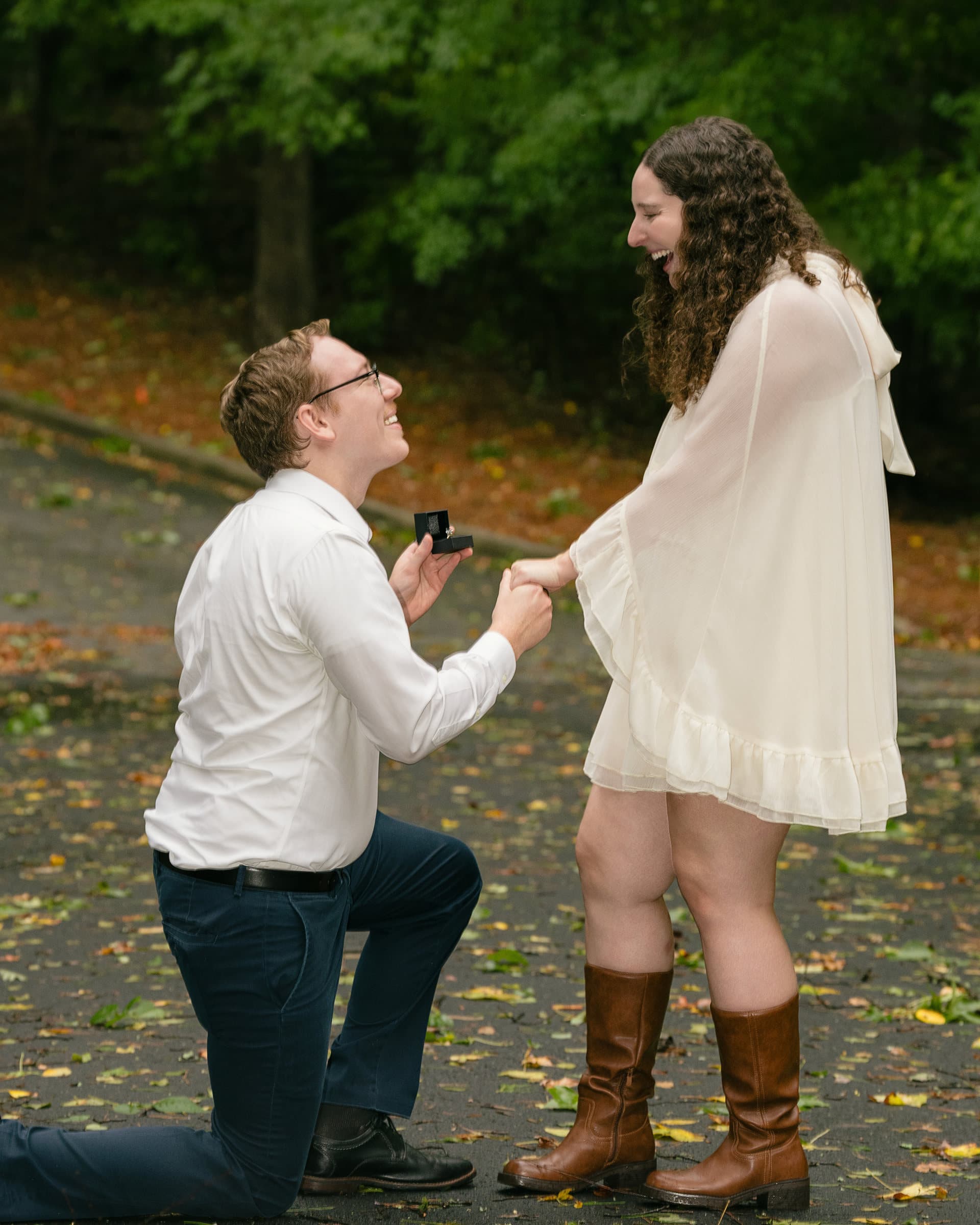 Luan on one knee asking the question, Sarah's hand to her mouth, rain-wet path
